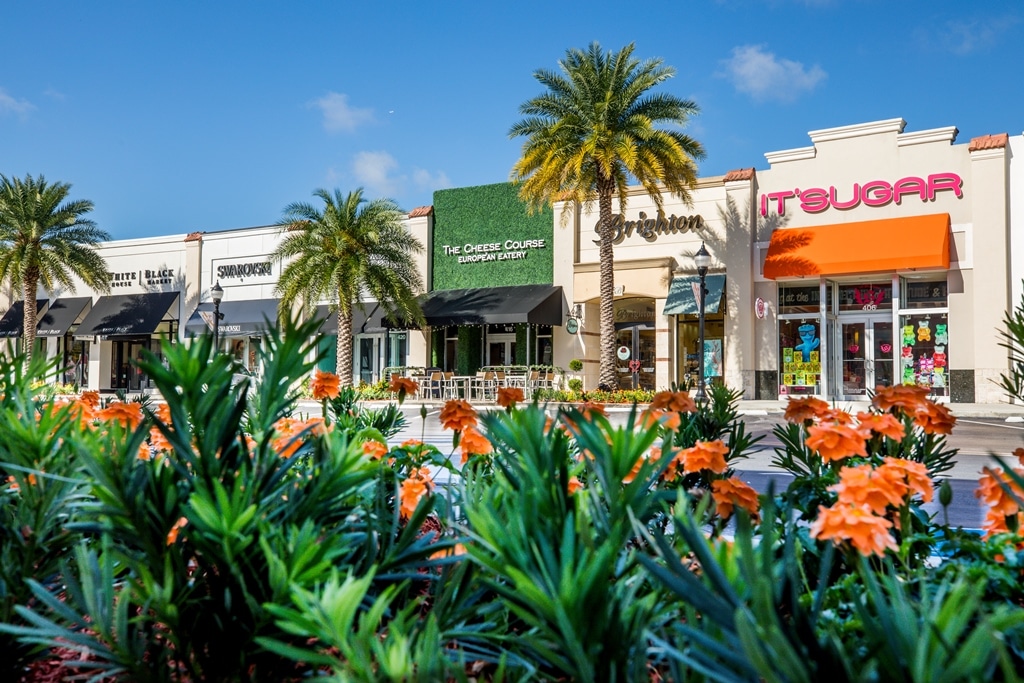 a row of storefronts at an outdoor shopping mall pictured from behind a colorfully landscaped traffic median