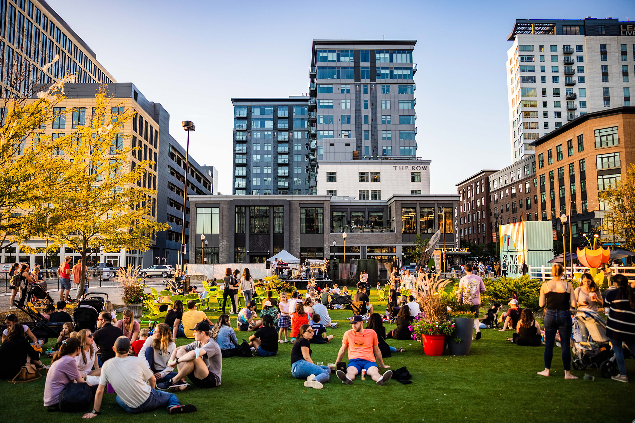 groups of picnickers gather for an event on a grass lawn in front of a mixed-use city backdrop