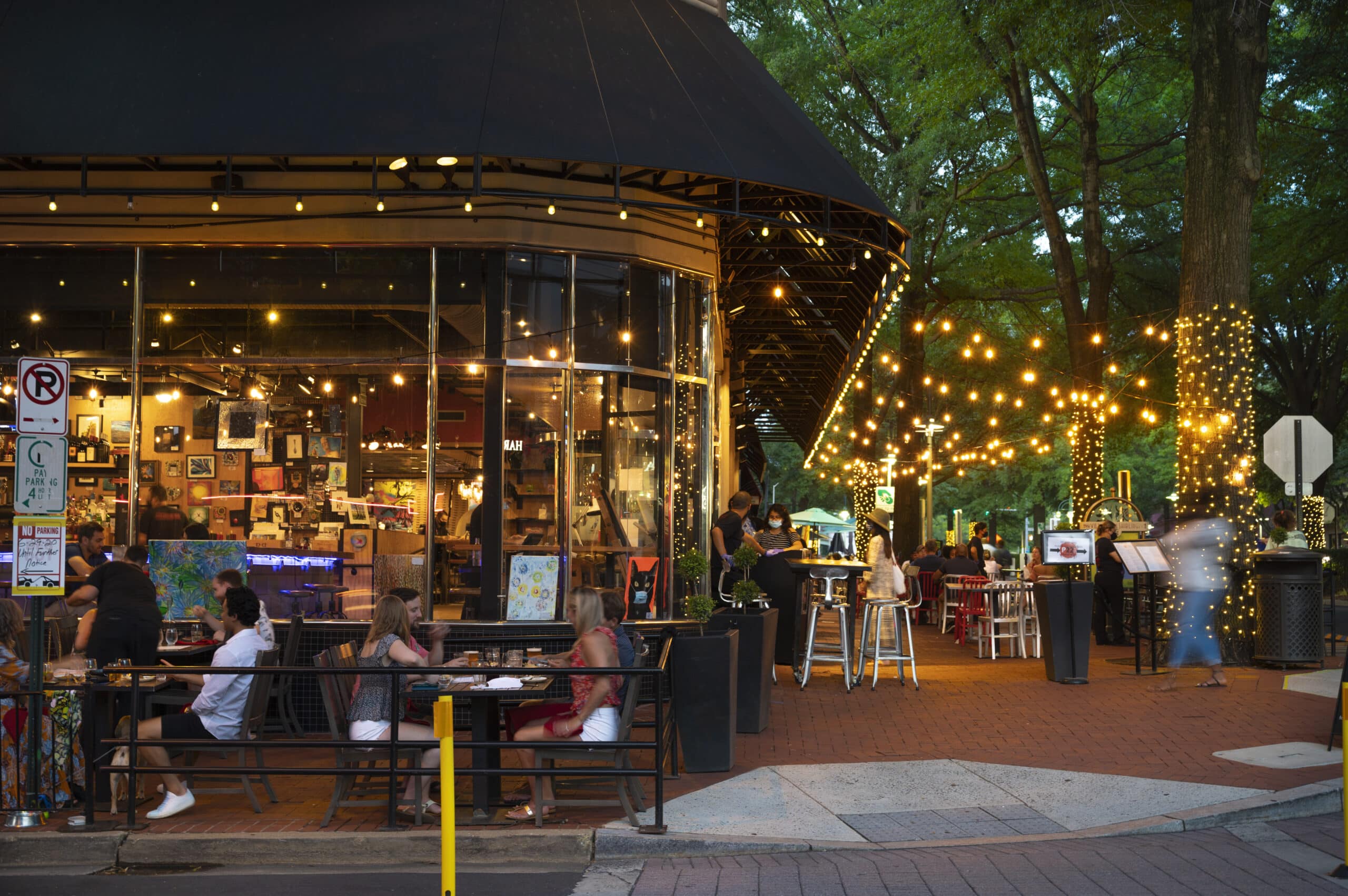 guests dine outside of a brightly-lit corner cafe at dusk with an assortment of artwork visible through cafe window